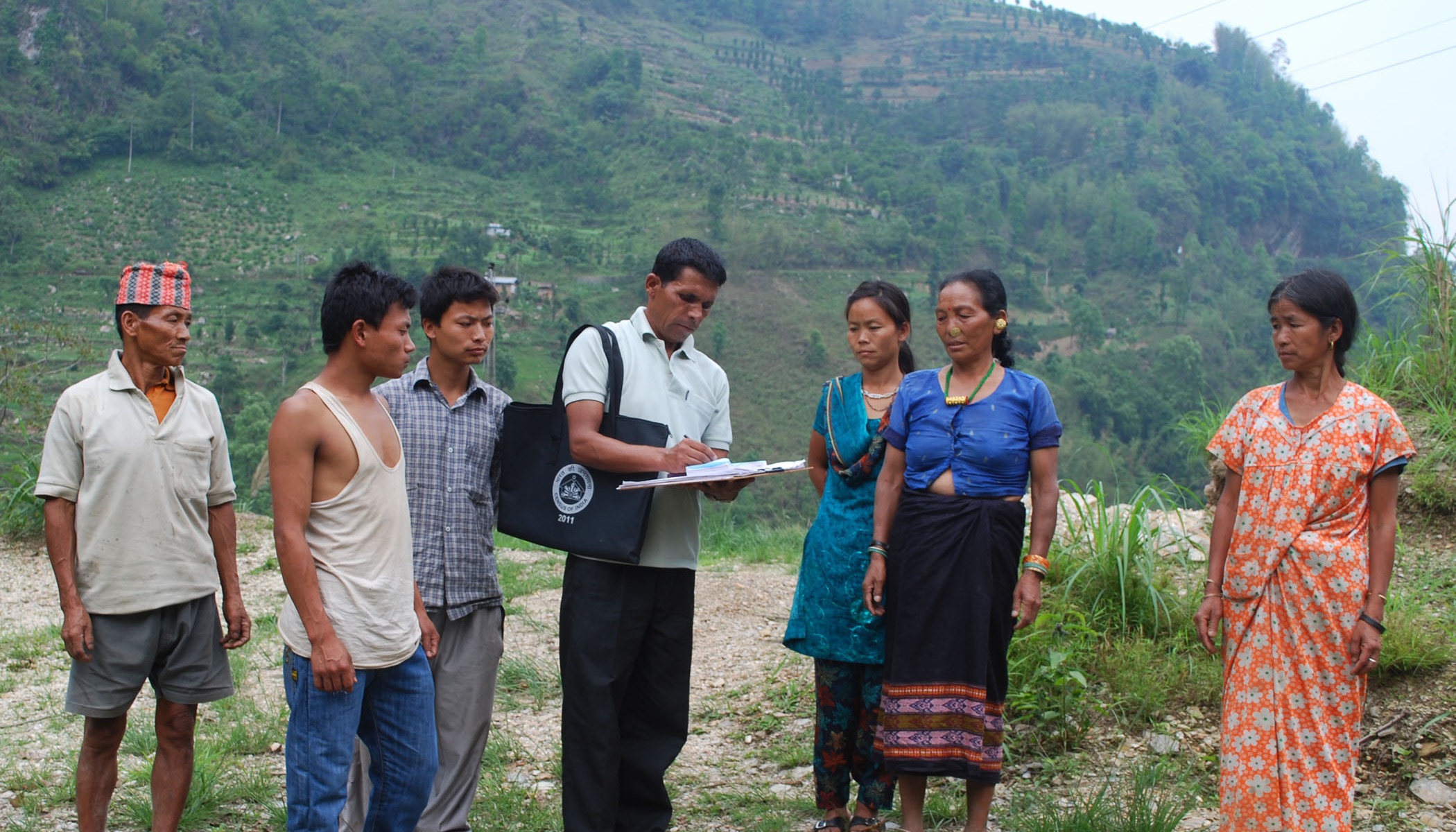 A Census of India 2011 enumerator, carrying an official Census of India 2011 bag, fills out a household schedule by hand while a group of villagers looks on, near Rinchengpong, West Sikkim